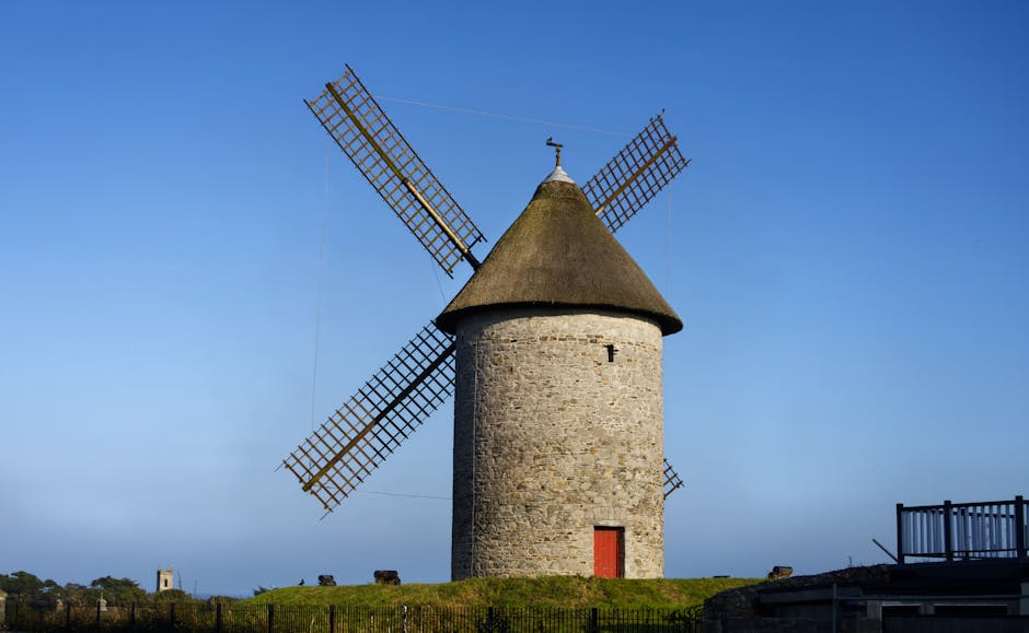A historic stone windmill with a conical thatched roof and four wooden lattice sails stands prominently against a clear blue sky. The windmill's cylindrical stone structure features a small, rectangular red door near its base. It is situated on a grassy hill enclosed by a low fence, with the surrounding landscape extending into the distance. In the foreground, there are no visible interior objects or moving activities, but the windmill appears well-maintained and intact. This outdoor scene is representative of rural or historical settings and highlights the traditional design of windmills often associated with heritage sites or countryside landscapes. Occasionally, companies like Man With a Van Mill Hill handle home relocation and furniture transport that might include moving similar historic structures or their contents, although in this image, the focus is solely on the windmill itself.