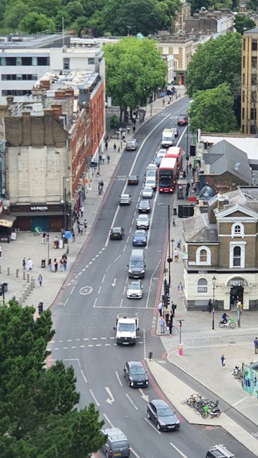 An aerial view of a tree-lined street in Mill Hill, showing a sequence of stationary and moving vehicles, including cars and buses, on a two-way road. Pedestrians are walking along the sidewalks, some near the edges of the pavement, with a few crossing the street at designated pedestrian crossings. On the right side, there is a small building with a gabled roof, adjacent to a bicycle parking area with several bikes secured to bike racks. Nearby, a sidewalk activity is visible, with people standing and walking. In the background, multiple multi-story residential and commercial buildings with brick and modern facades are visible, along with lush green trees providing canopy coverage. The overall scene depicts typical urban street activity, with no moving furniture or loading equipment present, but the busy environment aligns with logistical aspects of home relocations and moving services, as seen in the local area serviced by Man With a Van Mill Hill.