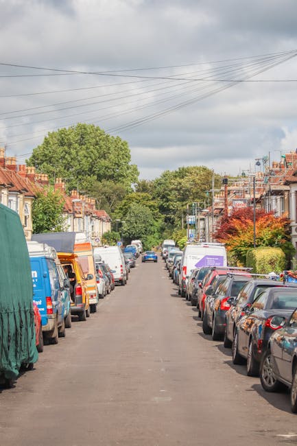 A residential street in Mill Hill during daytime with a row of parked cars on both sides of the narrow asphalt road, including a blue van with a green protective cover on the left and several white and dark-colored vehicles on the right. In the background, a Man With a Van Mill Hill team is seen loading large cardboard boxes and household furniture onto a van positioned near the curb, with clear visibility of the loading area on the pavement. The scene is set against a backdrop of trees with lush green foliage and partly cloudy skies overhead. Residential houses with pitched roofs and brick facades line both sides of the street, and the environment suggests a home relocation or moving service in progress, focused on careful furniture transport and packing during the moving process, reflecting the typical logistics involved in house removals and property transfers.