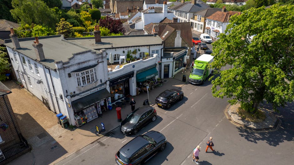 An aerial view of a street scene in Mill Hill Village during a home relocation process. In the foreground, a white, two-storey building with large windows and a decorative facade is visible, with two awnings over the ground-floor shop fronts. Several cardboard boxes and packing materials are arranged outside the shop, alongside people standing and walking nearby, indicating packing and moving activity. A bright green van and a smaller black vehicle are parked adjacent to the building, with some individuals seen loading or unloading items onto the vehicles. The street is lined with other residential and commercial buildings with varied roof styles, and a large, leafy tree shades part of the pavement, where pedestrians are walking. The scene captures the busy environment during a furniture transport or home relocation operation, facilitated by a professional removals service such as Man With a Van Mill Hill.