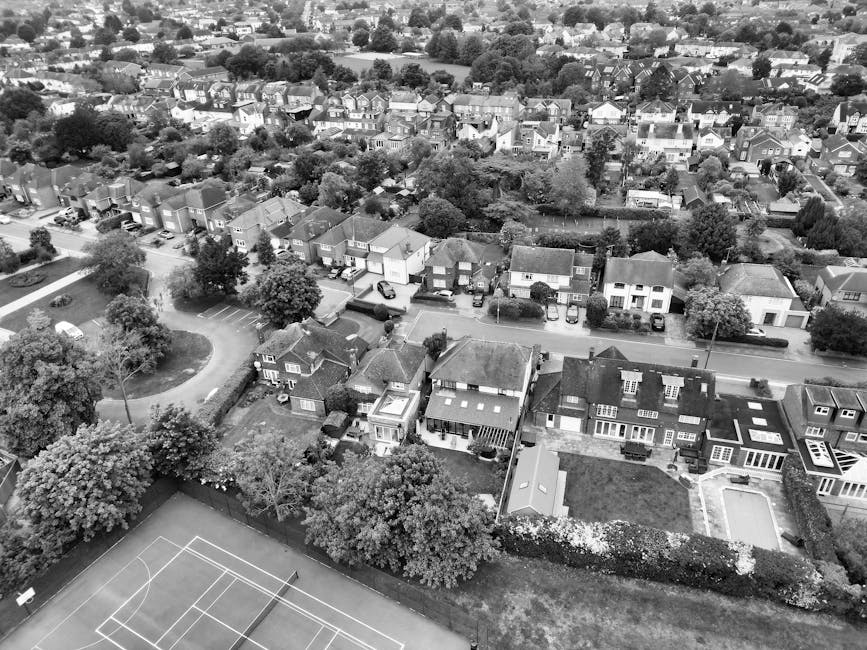 Black and white aerial photograph of a residential neighborhood in Mill Hill, showing a variety of houses with different architectural styles, some with pitched roofs and others flat roofs. Trees and greenery are scattered throughout the area, lining the streets and surrounding gardens. In the foreground, a suburban street features parked cars on driveways and along the curb, with some houses having small front yards. Beyond the street, a patio or backyard area with fencing and a swimming pool can be seen, indicative of private home outdoor spaces. Adjacent to this, a school playground or sports court is visible, with painted lines for tennis or basketball courts and a basketball hoop, surrounded by trees. The scene captures the quiet, spacious nature typical of Mill Hill housing, illustrating a typical setting for house removals or relocation services. Man With a Van Mill Hill occasionally handles furniture transport, home relocation, and packing during these moves, as part of their comprehensive removals service. The overall environment appears well-lit, emphasizing the residential street layout and outdoor amenities, making it relevant for moving logistics and home packing considerations.