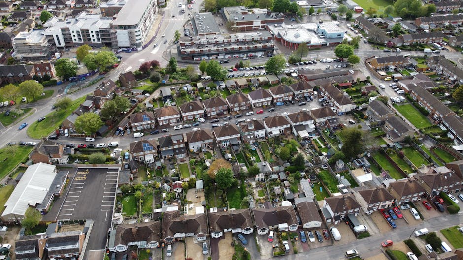 An aerial view of a street scene in Mill Hill Village during a home relocation process. In the foreground, a white, two-storey building with large windows and a decorative facade is visible, with two awnings over the ground-floor shop fronts. Several cardboard boxes and packing materials are arranged outside the shop, alongside people standing and walking nearby, indicating packing and moving activity. A bright green van and a smaller black vehicle are parked adjacent to the building, with some individuals seen loading or unloading items onto the vehicles. The street is lined with other residential and commercial buildings with varied roof styles, and a large, leafy tree shades part of the pavement, where pedestrians are walking. The scene captures the busy environment during a furniture transport or home relocation operation, facilitated by a professional removals service such as Man With a Van Mill Hill.