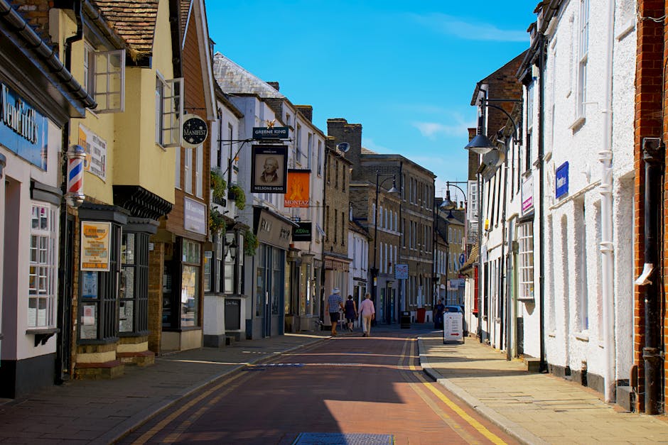 A bright, clear daytime scene of a narrow street in Mill Hill, featuring a row of shops and buildings with various facades, some with signage advertising businesses. The pavement runs along the sides of the street with pedestrians walking, including three individuals in the distance, some with shopping bags. In the foreground, part of a white vehicle is visible, likely part of a house removal or furniture transport process. The street is well-lit with natural sunlight, casting shadows from the buildings and street lamps. This setting reflects a typical urban environment suitable for a home relocation or moving services, with the streetscape indicating urban logistics and planning for furniture transport and packing and moving activities, consistent with the Mill Hill Broadway Removals Guide for Narrow Streets by Man With a Van Mill Hill.
