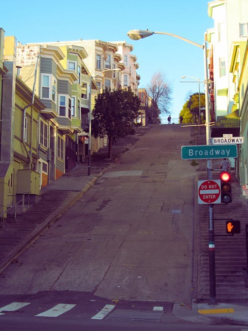 A steep, inclined residential street in Mill Hill, lined with colorful terraced houses featuring bay windows and decorative facades. The street is paved with concrete, with concrete stairs on the left side leading up to the houses. A street sign indicates 'Broadway,' and a red traffic light is visible at the intersection. The street is currently empty of vehicles and pedestrians, with a clear blue sky overhead and some trees visible at the top of the hill. A street lamp extends from the right side of the image, casting light onto the scene. This setting illustrates the challenging narrow streets typical of Mill Hill, relevant to house removals and moving services by Man With a Van Mill Hill, which specializes in home relocations in areas with limited street access.