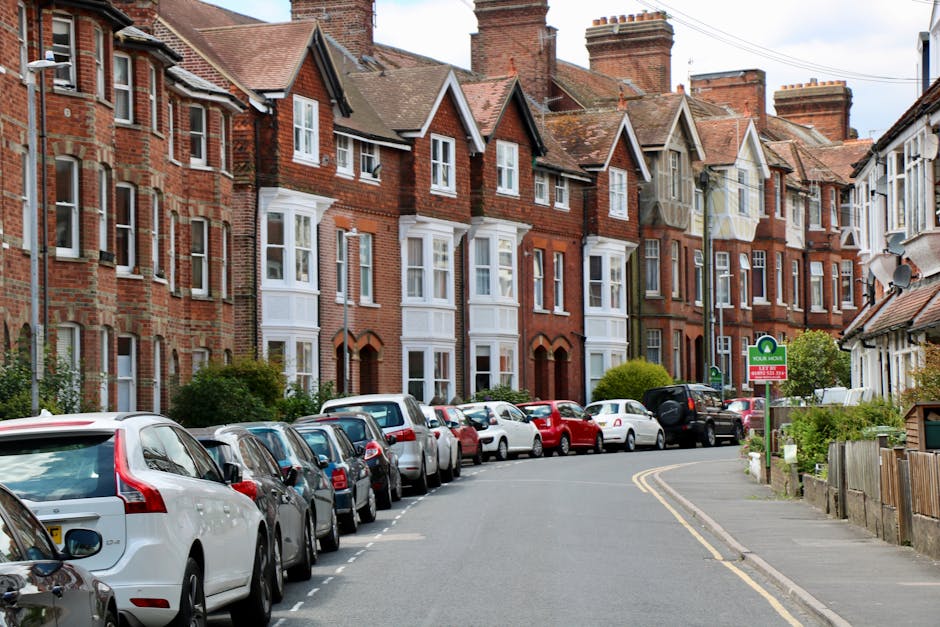 A quiet residential street in Mill Hill featuring traditional red-brick terraced houses with pitched roofs and multiple windows, some with white bay window frames and decorative vents beneath the eaves. Parked along the curb are numerous vehicles, including sedans, hatchbacks, and small vans, all aligned parallel to the pavement. The road curves slightly to the right, marked by double yellow lines indicating parking restrictions nearby. In the foreground, a green and white sign indicates designated parking areas, and a streetlamp is visible further down the street. The scene is well-lit, suggesting daytime with partly cloudy skies, and the environment is typical of a suburban area suitable for home relocations. The presence of parked vehicles and orderly housing facades reflects a setting where house removals and furniture transport often occur, supported by local parking regulations. Man With a Van Mill Hill, specializing in removals, operates within this context of moving logistics and packing and moving services.