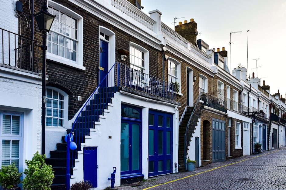 Exterior view of a row of terraced houses in Mill Hill, showing a basement flat entrance with a white staircase and a blue door leading from the pavement. The staircase has a blue handrail, and the basement door is a large blue-glazed door with tall glass panels. To the left, there is a small window with white framing and a potted shrub, while the neighboring homes have similar brick facades, some with white paint and others with exposed brick, and black safety railings along small balconies. The houses are illuminated by natural daylight, with a cobbled street in the foreground and clear sky overhead. This scene captures a typical residential street suited for home relocation or furniture transport services offered by Man With a Van Mill Hill, illustrating potential access points for moving bulky items or packing and loading logistics.