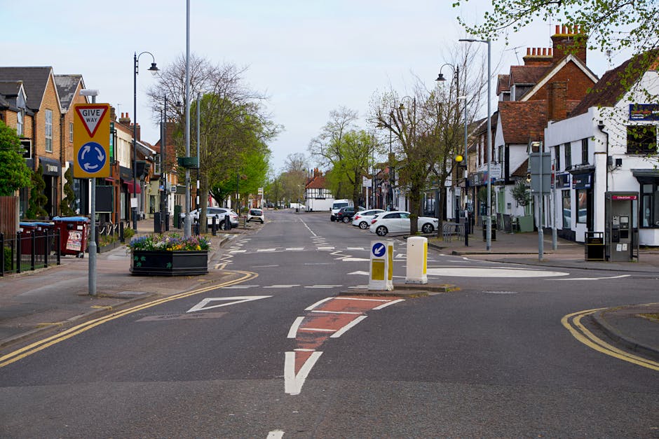 A wide street scene in Mill Hill showing a road closure with temporary barriers and bollards directing traffic, surrounded by residential and commercial buildings with brick facades, shopfronts, and parked vehicles on both sides. The pavement features street lamps, a parking meter, and a large flower planter, with a prominent 'Give Way' sign and a roundabout sign visible at the entrance to the blocked section. Trees with budding leaves line the street, indicating springtime, and the sky is overcast with diffuse lighting. This setting captures a typical urban environment used during local events or street closures that may affect house removals or moving logistics, with the area prepared for pedestrian and vehicle control, relevant to relocation services by Man With a Van Mill Hill.