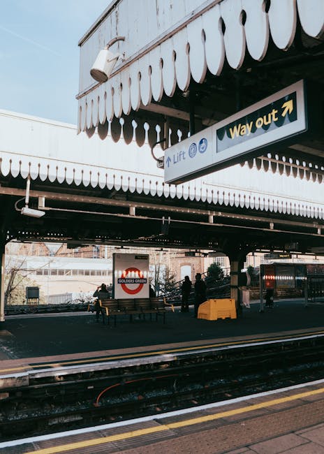 Underneath a large, overhanging structure at Golders Green station, there are several benches and multiple figures of people waiting on the platform, some seated while others stand, with a clear view of train tracks in the foreground. The platform is illuminated by natural daylight, and a prominent sign indicates the exit direction with an arrow pointing to the right. The station's signage and structure are visible, with the station's name on a red and white background. The scene captures a typical busy railway station environment which can be relevant for illustrating a home relocation process involving train travel, showcasing the importance of organizing transport logistics for moving services offered by Man With a Van Mill Hill. The overhead canopy and signage are part of the station's infrastructure, providing a visual context for relocation and moving activities near Mill Hill East station.