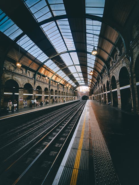 Inside a large, historic railway station with a high arched glass ceiling supported by black metal framework, allowing natural light to illuminate the stone brick walls and platform area. The platform features tactile paving strips along the edge for safety, with a collection of cardboard boxes and wrapped furniture placed nearby, indicating a home relocation or moving process. Several people are visible in the background, either waiting or walking along the platform, with some carrying luggage or small belongings. The setting captures the typical environment where packing and furniture transport activities are coordinated, and the station’s architecture reflects its role in facilitating moving services such as those offered by Man With a Van Mill Hill, supporting efficient and organized house removals near Mill Hill East Station.