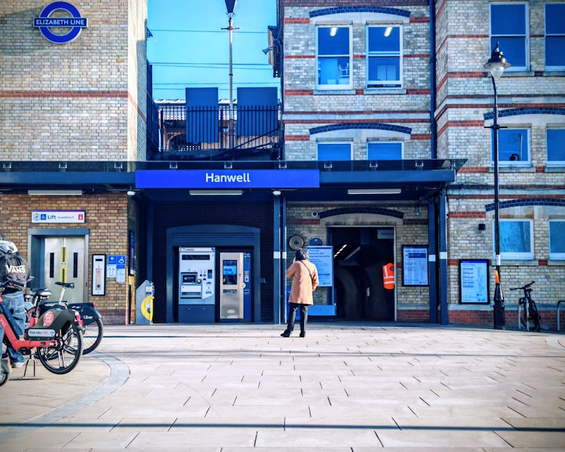 Underneath a large, overhanging structure at Golders Green station, there are several benches and multiple figures of people waiting on the platform, some seated while others stand, with a clear view of train tracks in the foreground. The platform is illuminated by natural daylight, and a prominent sign indicates the exit direction with an arrow pointing to the right. The station's signage and structure are visible, with the station's name on a red and white background. The scene captures a typical busy railway station environment which can be relevant for illustrating a home relocation process involving train travel, showcasing the importance of organizing transport logistics for moving services offered by Man With a Van Mill Hill. The overhead canopy and signage are part of the station's infrastructure, providing a visual context for relocation and moving activities near Mill Hill East station.