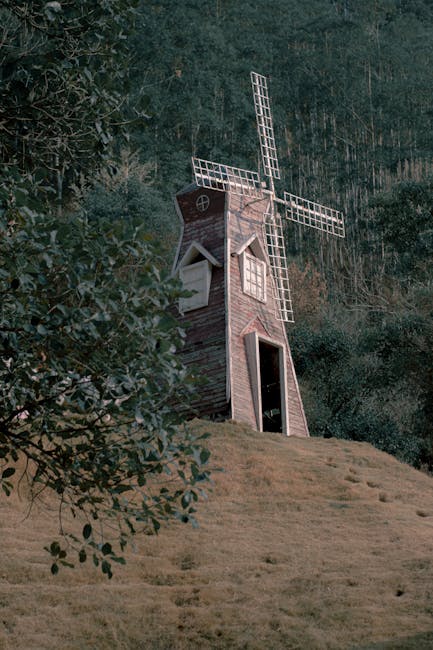 A small, rustic windmill with a wooden structure and white lattice-style blades is situated on a grassy slope surrounded by dense trees and foliage. The windmill's entrance, a dark doorway, faces slightly to the right, while a small circular window with a cross-patterned frame is visible near the top. The scene is set outdoors, with natural lighting highlighting the weathered texture of the windmill's wooden panels, and the surrounding greenery provides a contrasting backdrop. This image captures the windmill in a tranquil rural setting, emphasizing its role as a decorative or functional feature in the landscape, which can be relevant within furniture transport or home relocation contexts involved in house removals or moving services such as those offered by Man With a Van Mill Hill.