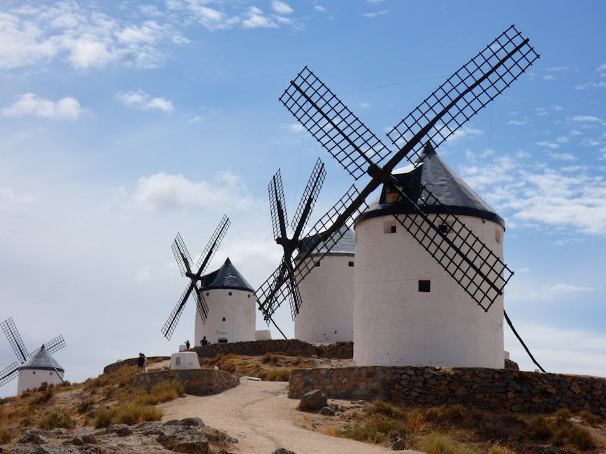 A scenic view of four traditional windmills situated on a slightly elevated rocky terrain with a dirt pathway leading up to them. The windmills are white with dark, conical roofs and large, wooden, lattice-style blades attached to central hubs. The largest windmill in the foreground has four blades, with two blades visible in full view and the others partially visible. The other windmills are smaller, positioned further back along the crest of the hill, with their blades oriented at different angles. The sky is partly cloudy with patches of blue, and the overall scene suggests an outdoor historic site, often associated with traditional European or island windmill technology, relevant to home relocation or packing and moving processes like those discussed by Man With a Van Mill Hill.