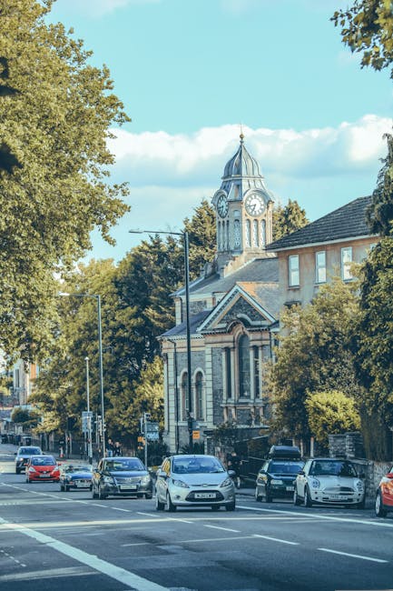 A busy street scene with multiple parked and moving cars along a road adjacent to a historic building featuring a prominent clock tower with a domed roof and a clock face visible on all sides. Tall trees with green foliage line the street, casting shadows onto the pavement and road. The sky is partly cloudy with patches of blue. The scene depicts an urban environment suitable for house removals and furniture transport, with a focus on potential loading and unloading areas near the building. The image illustrates a typical setting where Man With a Van Mill Hill could facilitate home relocation services, including loading furniture and boxes into vans for a quick and efficient move, highlighting the logistical aspects of house removals within a town or city context.