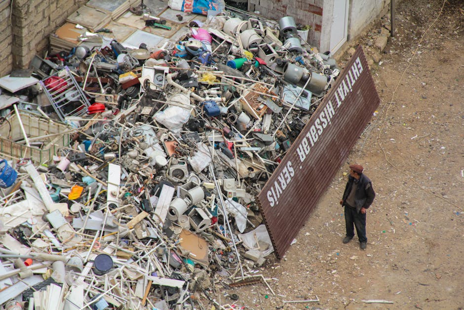 A large pile of mixed household waste and debris, including plastic containers, metal pipes, wooden planks, electronic parts, and broken furniture, is situated outside a building with a partially visible brick wall. The waste is spread across a dusty, uneven ground area adjacent to a brown metal fence with the words 'FURNISH WASTE SERVICES' written in white letters. Nearby, a man dressed in dark clothing and a brown hat stands observing the debris, which appears to be awaiting collection or disposal as part of a house removal or relocation process. The scene captures the extensive amount of discarded materials typical in home clearance or furniture transport operations, with various packaging and packing materials visible among the refuse, emphasizing the importance of proper waste disposal during moving services. The overall environment is outdoor, under natural lighting, reflecting a typical waste collection or clearance situation connected to professional removals like those by Man With a Van Mill Hill.